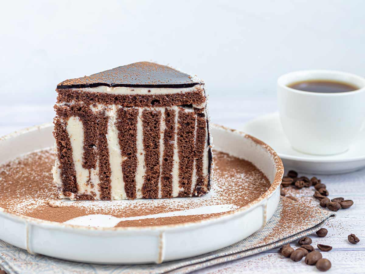 A slice of zebra cake with chocolate frosting and a cup of coffee beside the plate.