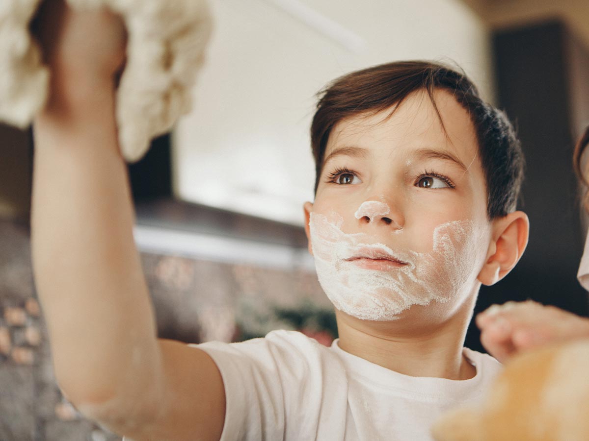 Young boy holding dough in the air with a beard of flour on his face.