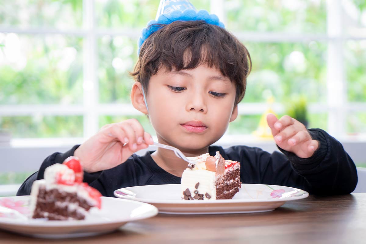 Young boy wearing a blue party hat sat at the table eating a piece of cake.