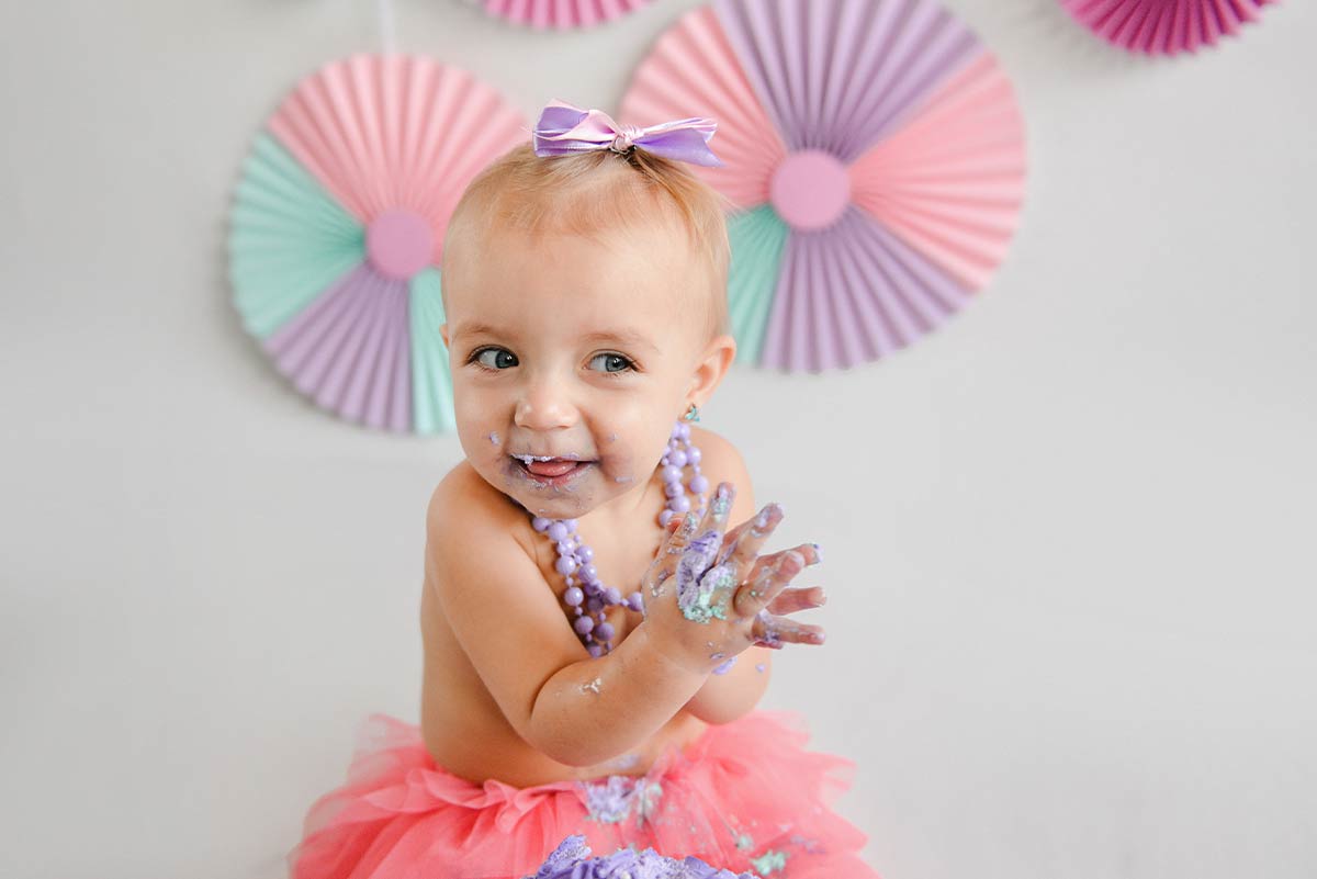 Baby girl wearing a tutu and necklaces with icing from a ninja turtle cake on her mouth and hands.