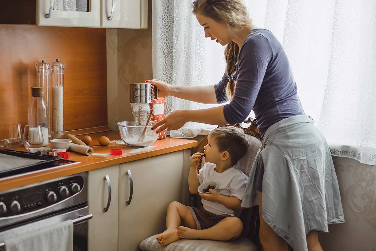 Mum in the kitchen baking a cake with her young son sat on the chair watching her.