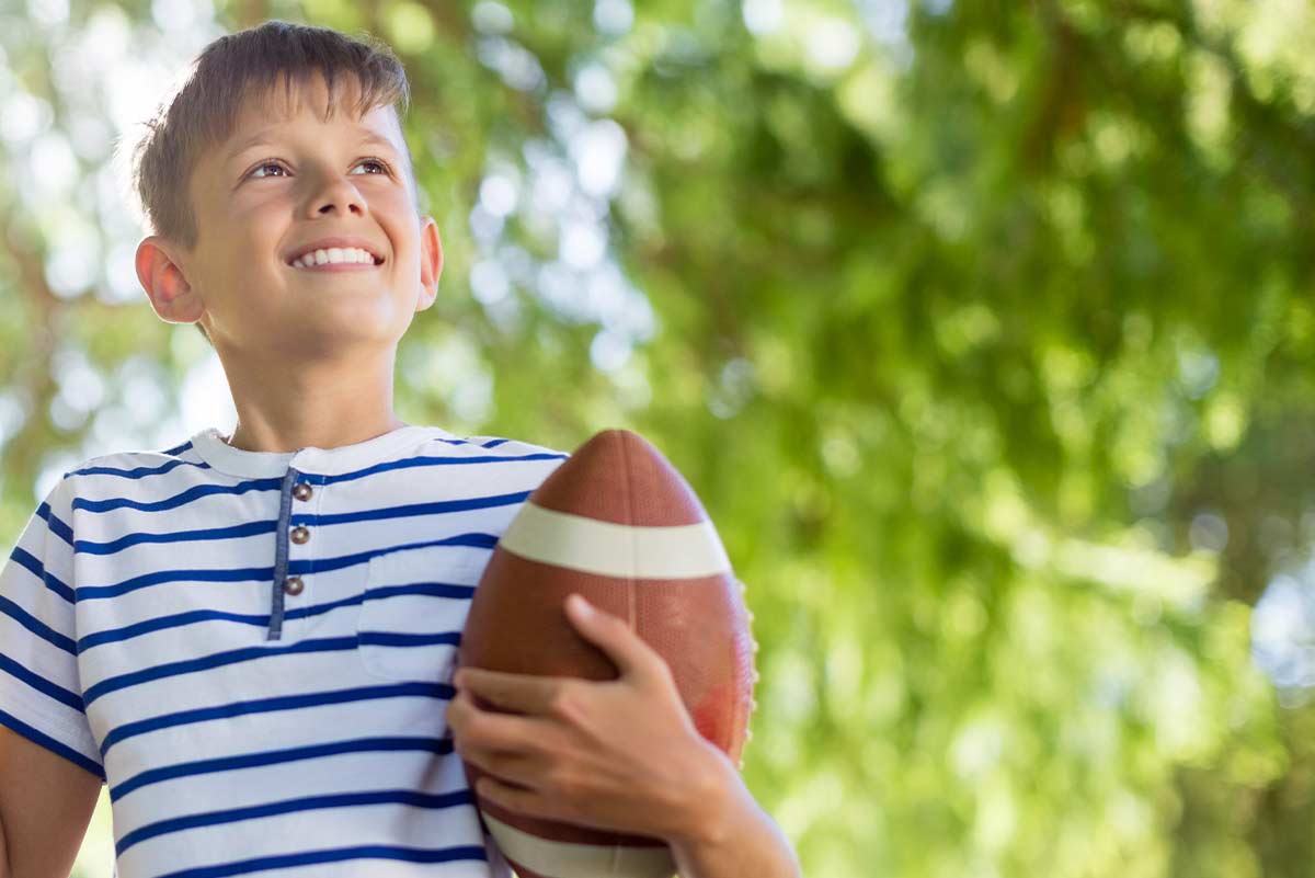 Young boy holding a rugby ball outside, smiling.