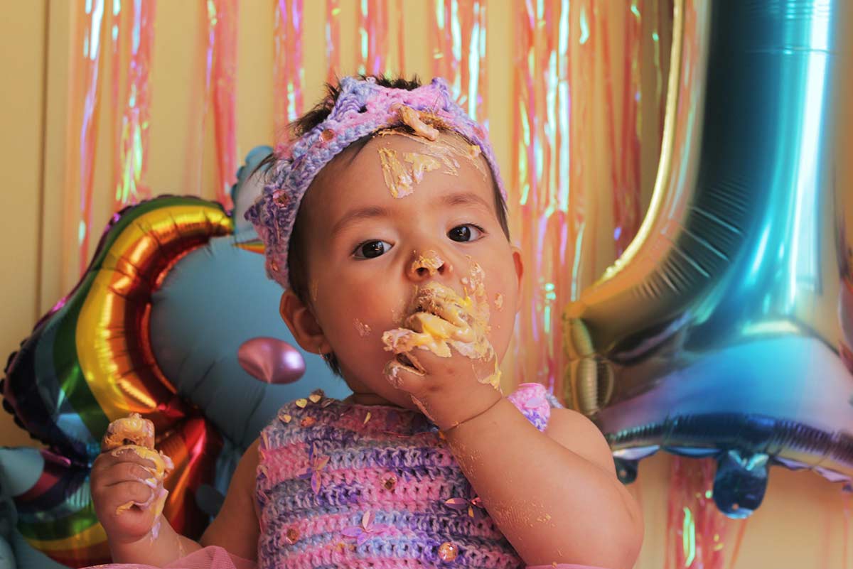 Toddler eating cake with her hands, icing all over her face and fingers.