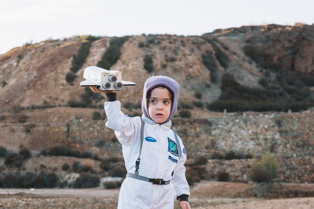 Child dressed as an astronaut standing outside holding up a toy rocket as if it's flying.