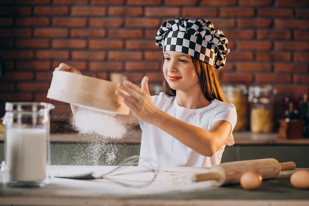 Teenage girl wearing a chef's hat, sifting flour in the kitchen to make a laptop cake.