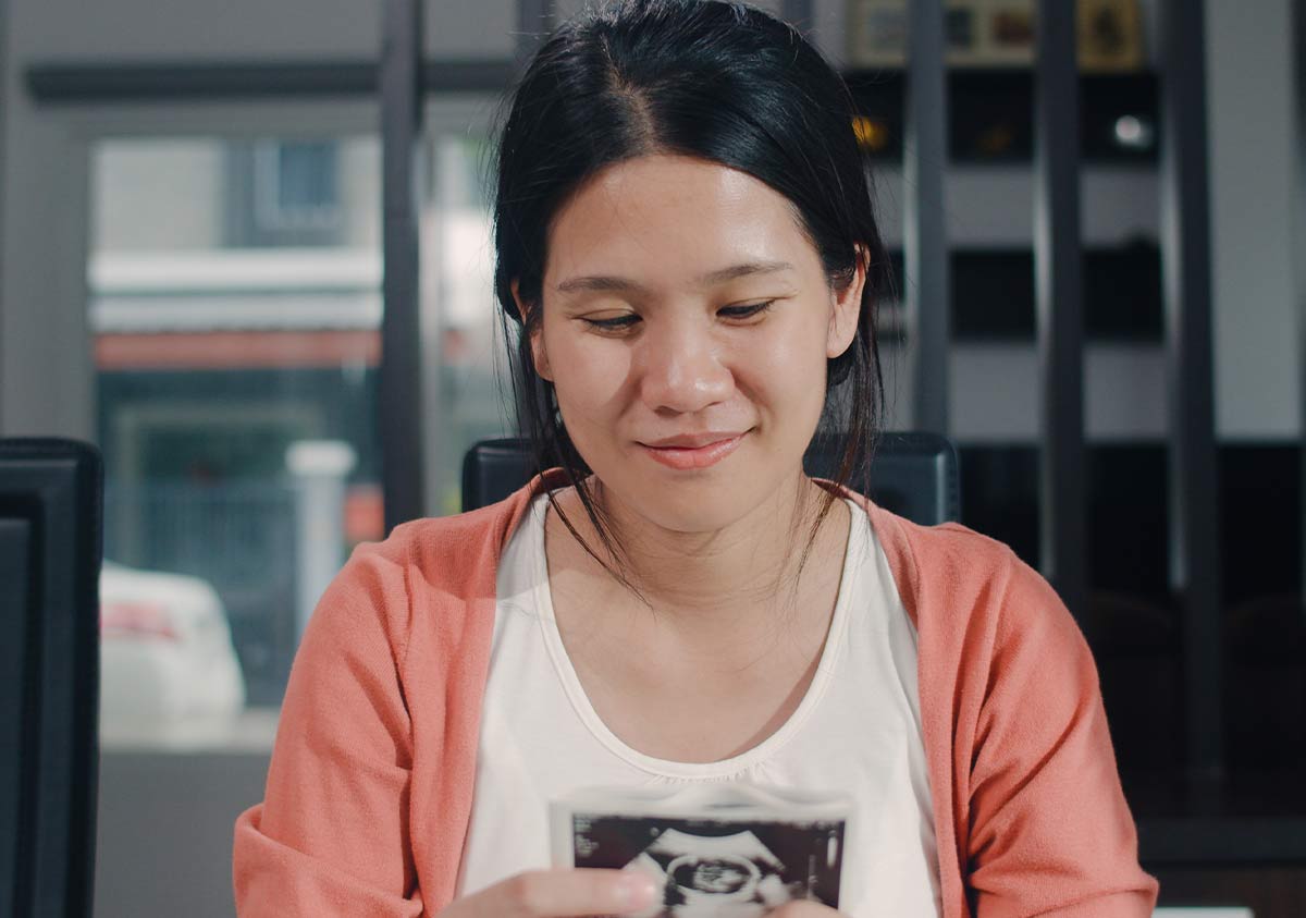 Woman sitting down smiling at a baby scan photo that she is holding.