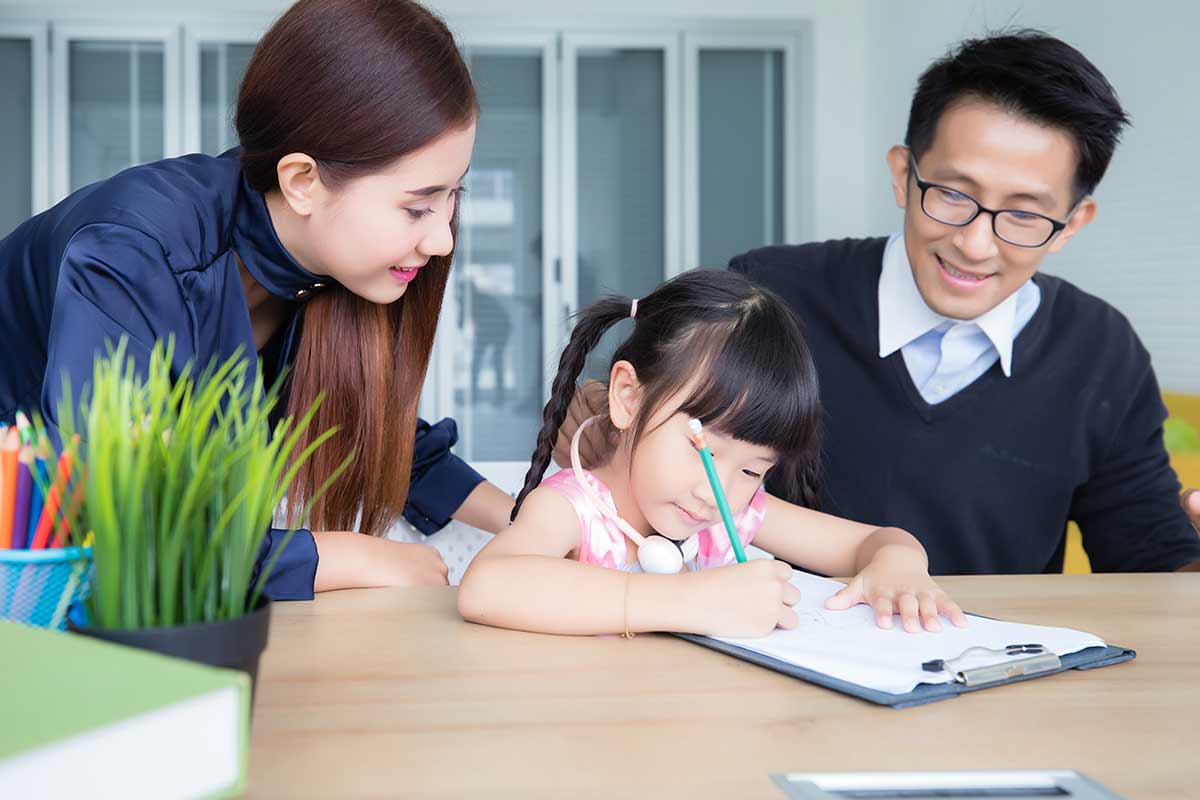 A young girl is sat at the table studying idioms whilst her parents, sat either side of her, watch proudly.