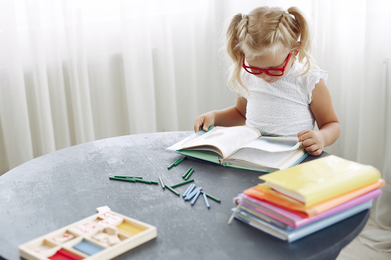 A young girl wearing red classes is sitting at the table, surrounded by colourful books, learning about idioms.