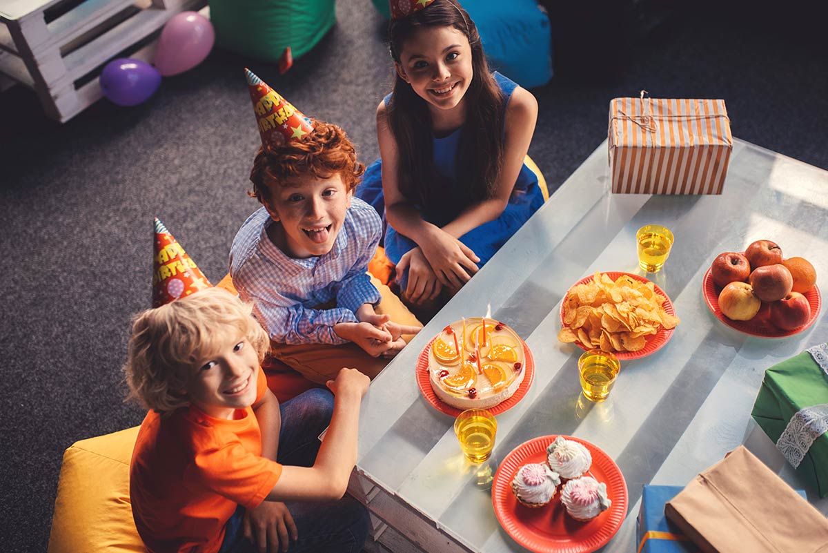 Three kids wearing party hats at around a table smiling and waiting to eat cake.