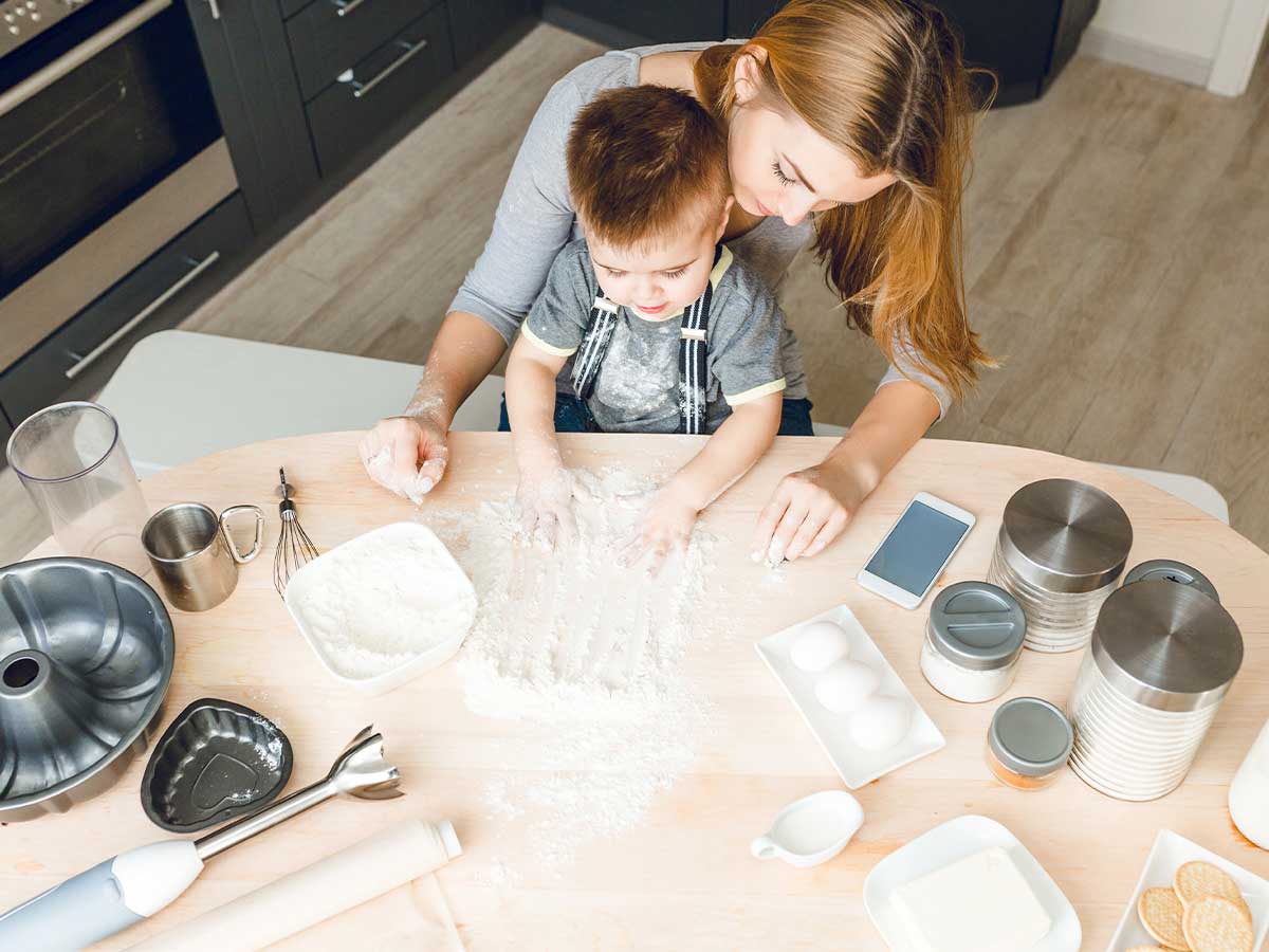 View from above of a young boy surrounded by baking equipment, helping his mother to prepare the flour to bake a LEGO cake.