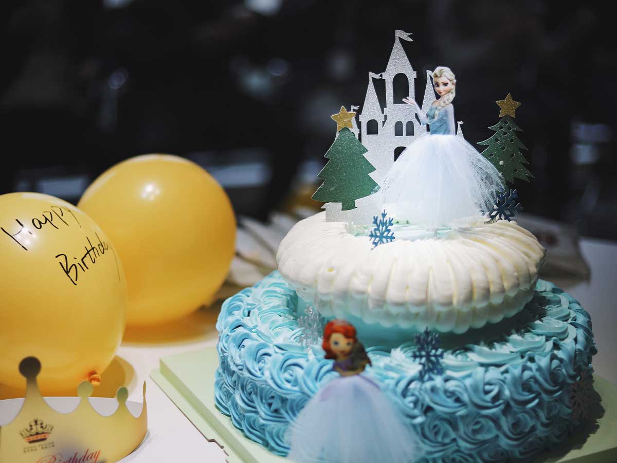 White and blue Frozen themed princess castle cake with Elsa and Anna cake toppers next to some birthday balloons.