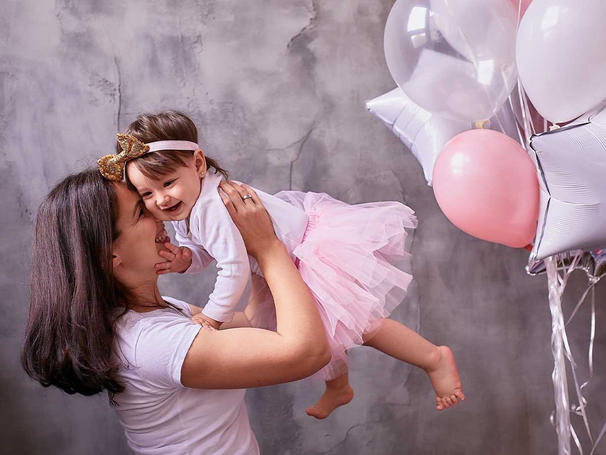 A mother smiles at her daughter as she lifts the toddler above her at a princess party next to some pink balloons.