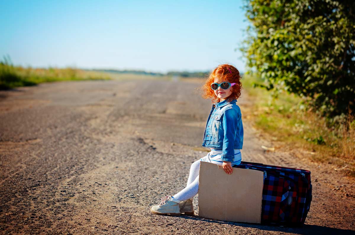 Toddler wearing sunglasses sitting on a suitcase on a country road looking happy everything has been packed for holiday.