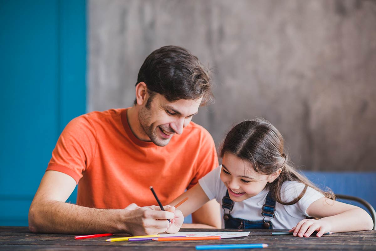Dad sat at table with daughter teaching her to tell time, both of them smiling.