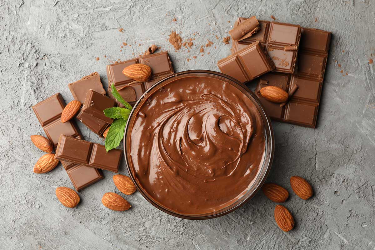 A bowl of melted chocolate surrounded by nuts and squares of chocolate that could be used to decorate a gravity cake.