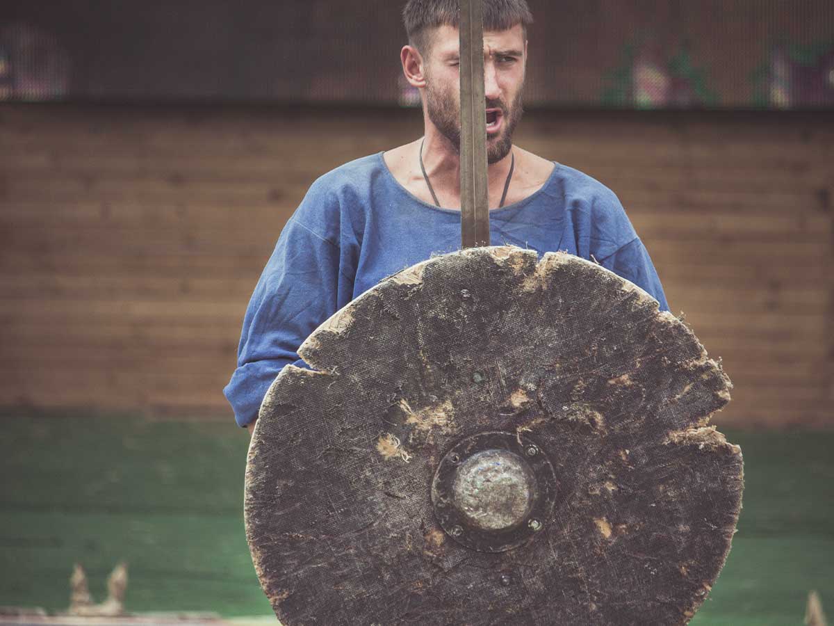 A man dressed in a blue cloth shirt holding a sword and a Viking shield.