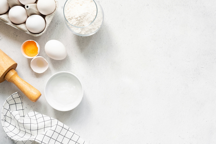 Ingredients needed to make a butterfly cake, including eggs, sugar and flour, laid out on a white table with a rolling pin and a tea towel.