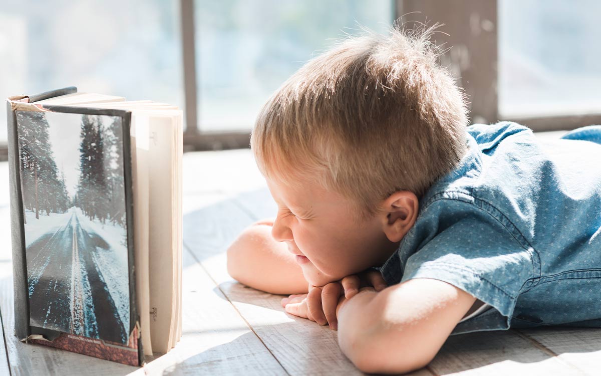 Young boy lying on his tummy on the floor squinting as he tries to read a book in the sunlight.