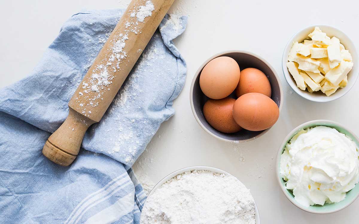 Some ingredients needed to make a superman cake, including eggs, butter and flour, are laid out on a white table with a rolling pin and a blue tea towel.