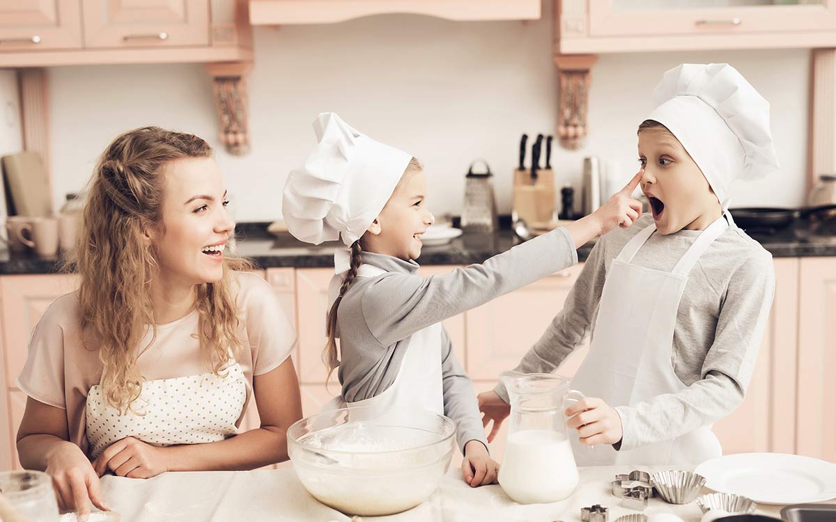 Three kids are smiling as they bake a superman cake together, one girl gently wipes flour on the boy's nose.