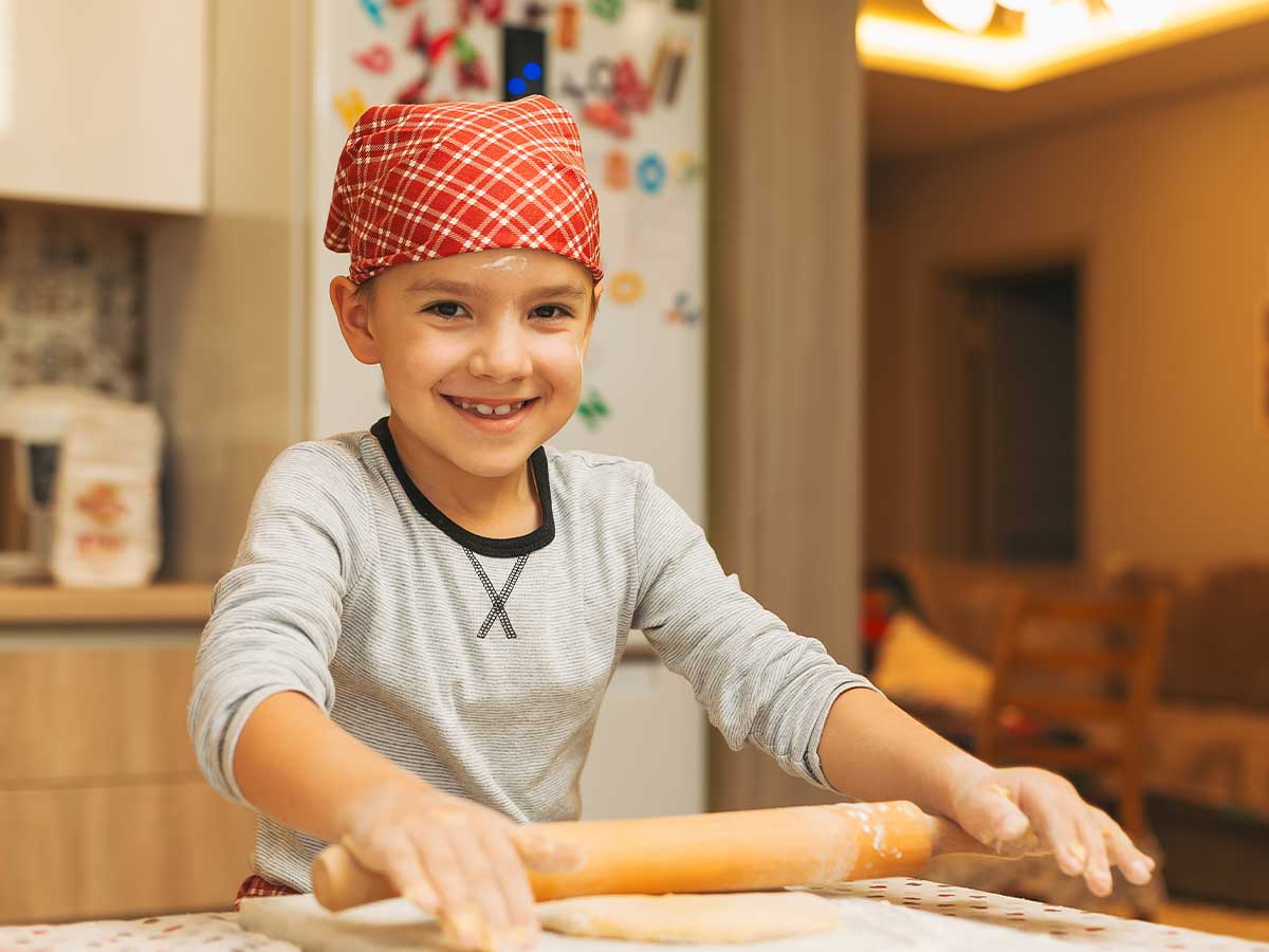 A young boy smiles at the camera as he uses a rolling pin to roll out fondant icing to decorate an Xbox cake.