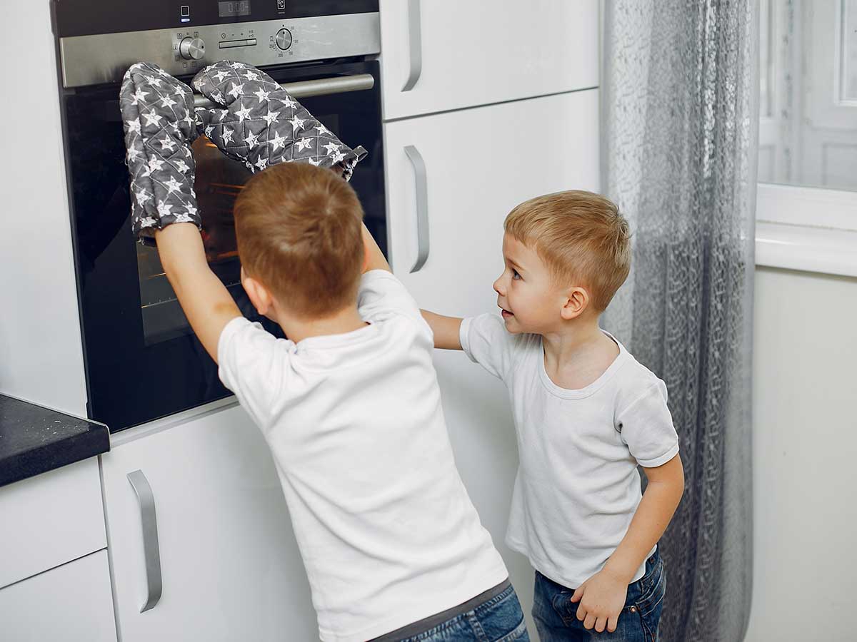 Two young boys, one wearing oven gloves, look into the oven to check on their Xbox cake.