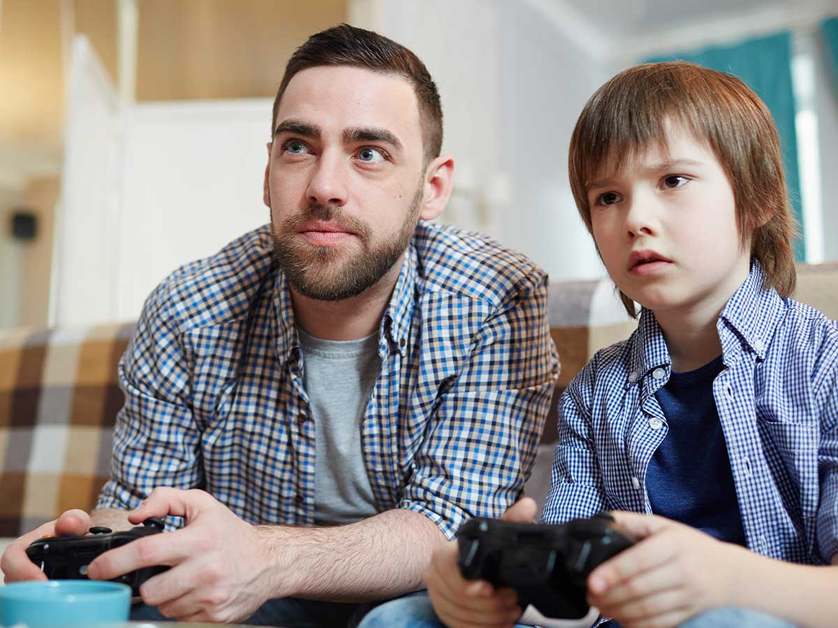 A father and son are sitting next to each other holding Xbox controllers, they both have a look of concentration on their face as they are playing an Xbox game.