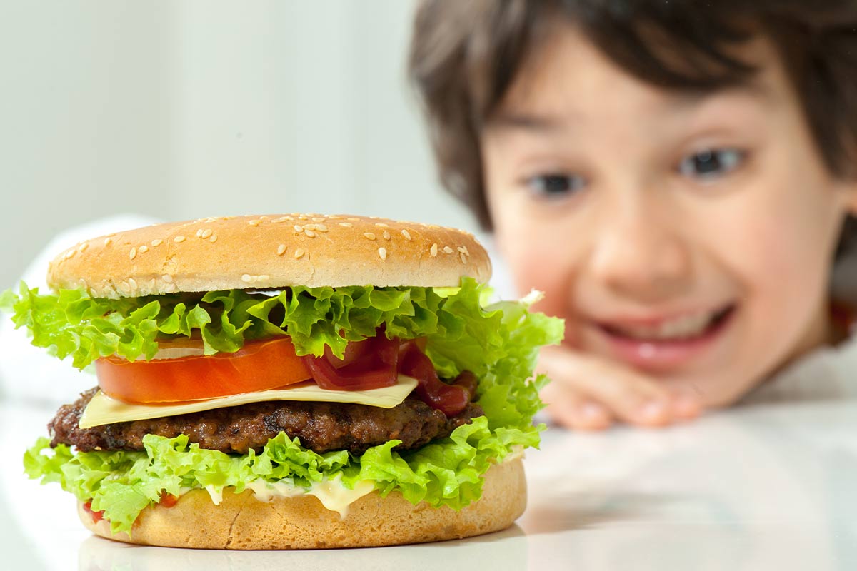 A burger is on the table in the foreground, behind it a young boy is smiling and looking at the burger.