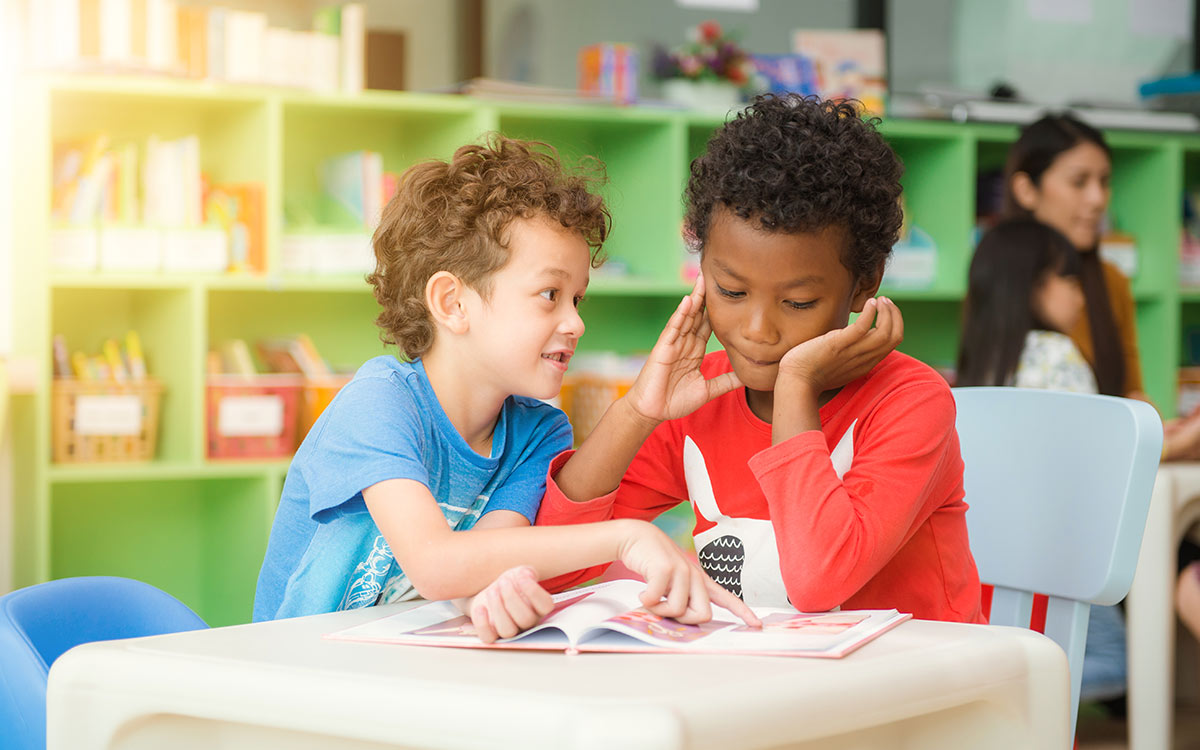 Two schoolboys are sitting at a table together looking at a book, they are learning how to use the present perfect tense.