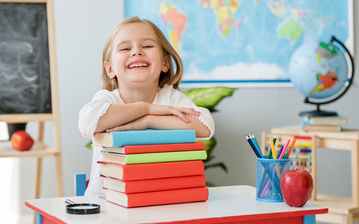 Girl standing in a classroom resting her arms on a pile of books smiling.