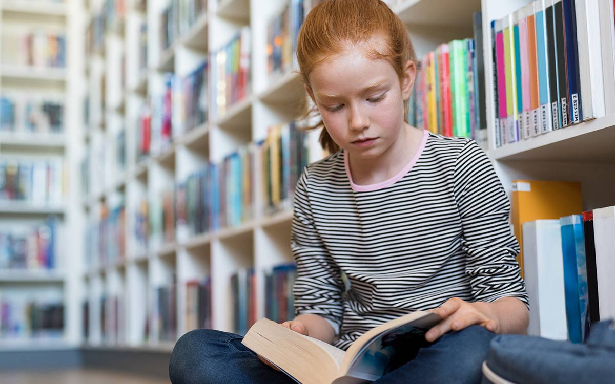 Young girl in KS2 sat on the floor in the library reading a book to help her recognise figurative language.r