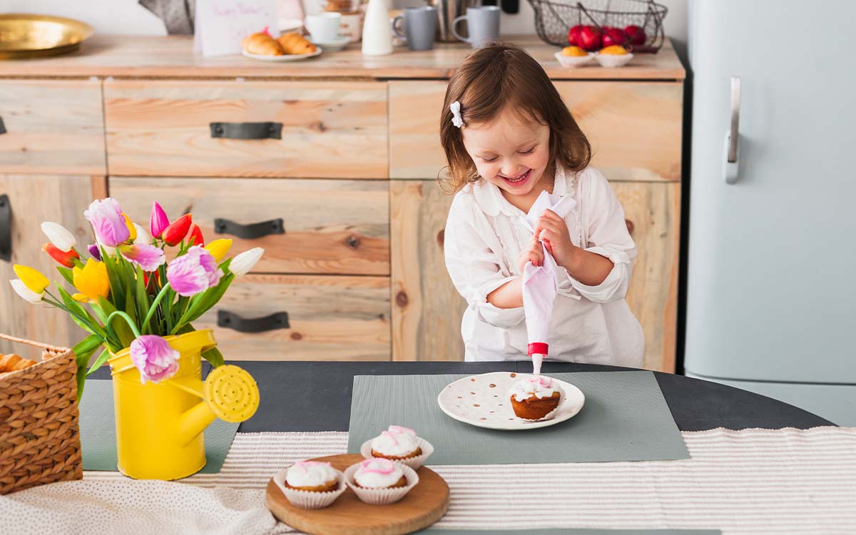 A young girl smiles as she uses a piping bag to ice some cupcakes.