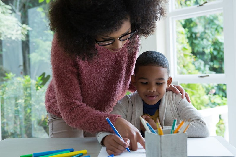 A mother leans over and helps her son to complete a WW2 art KS2 activity, he is using felt tips to draw on a piece of white paper.