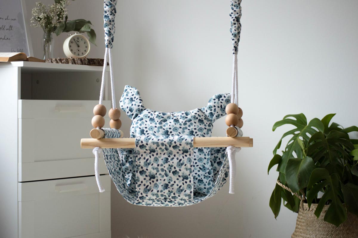 A blue and white baby swing hanging by white rope from the ceiling in a bedroom, next to a chest of drawers and a plant.h