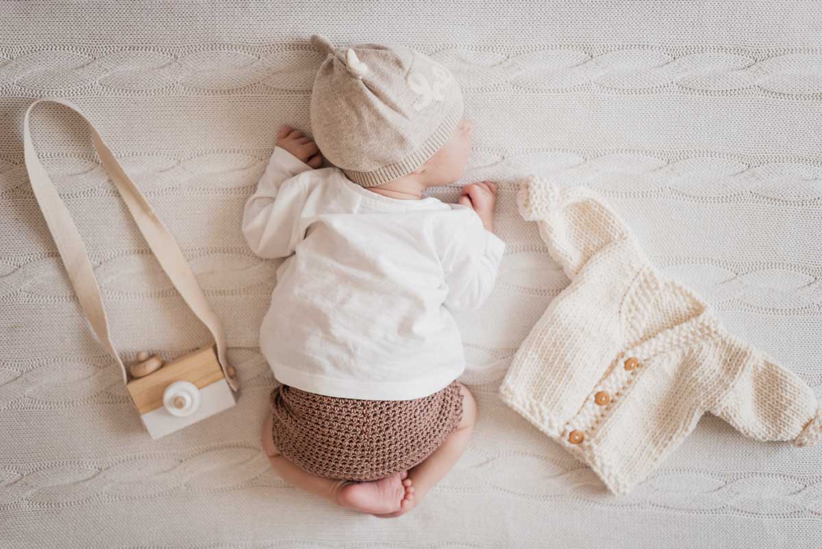 A baby wearing neutral colours is curled up on a blanket next to a toy camera and a knitted cardigan.