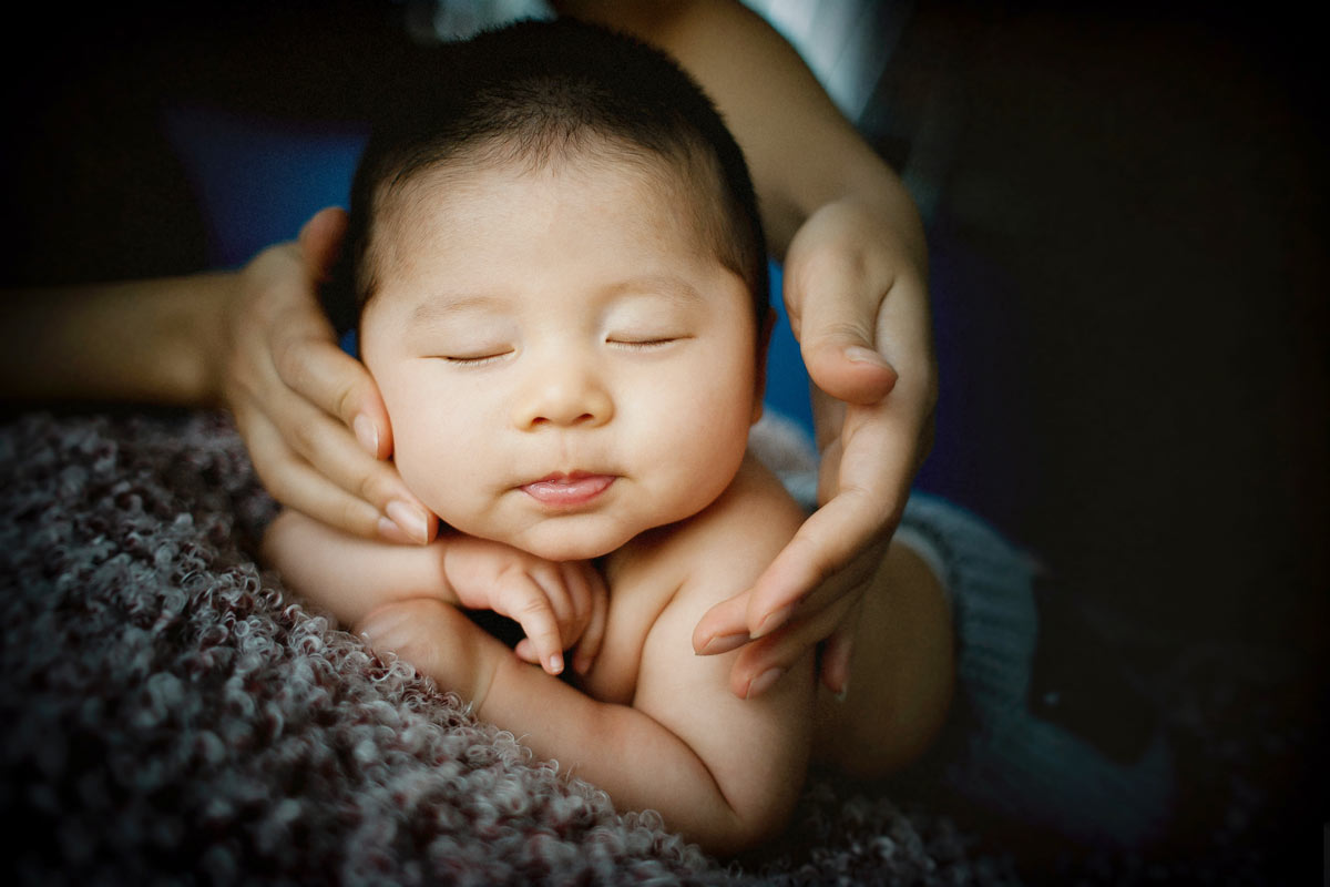 A newborn baby with his eyes closed lies on his front and rests his arms on a cushion whilst an adult's hands support his head.