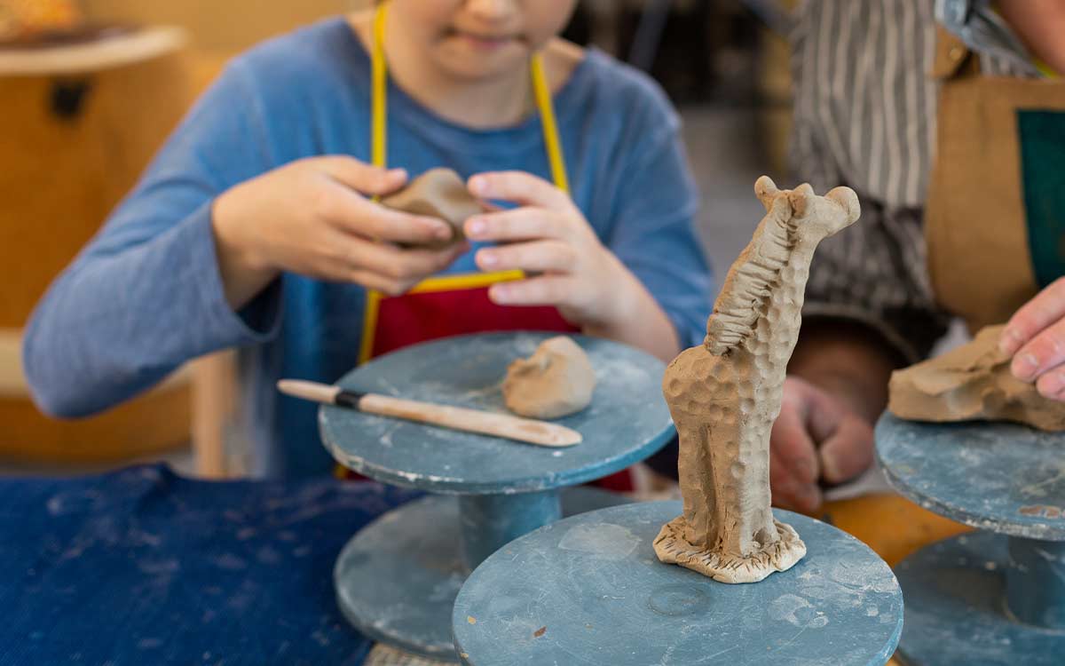 Close up image of a model giraffe, in the background a child uses clay to make a model giraffe craft.
