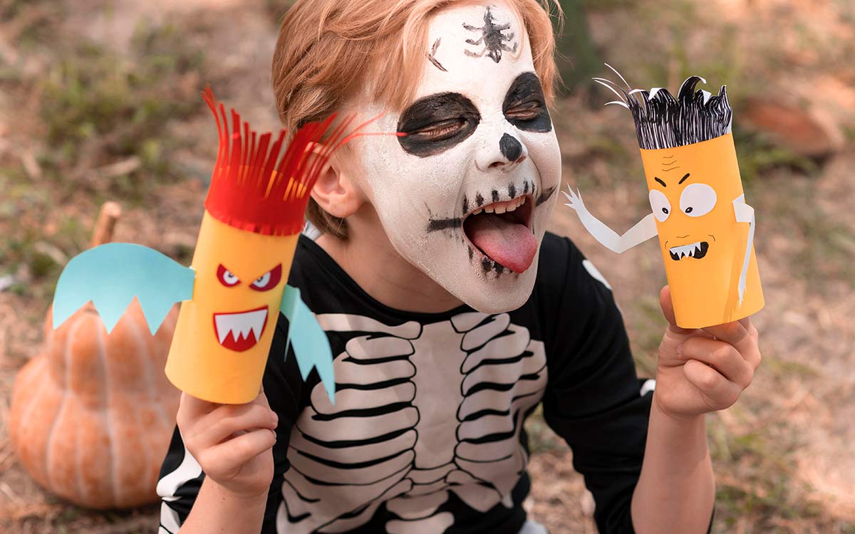A young boy dressed as a Halloween skeleton with skeleton face paint sticks his tongue out whilst holding some Halloween crafts.