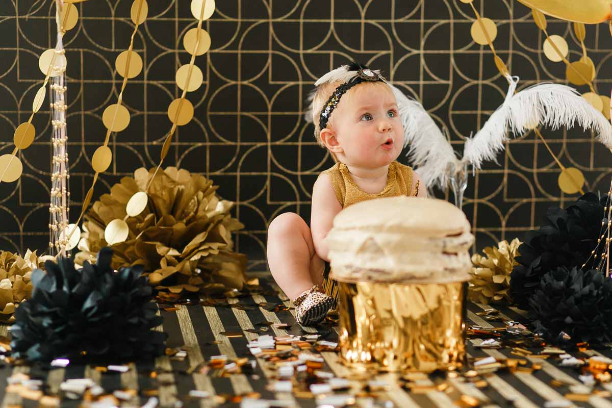 Baby girl wearing a gold dress sat on the floor in a black and gold room with gold decorations everywhere.