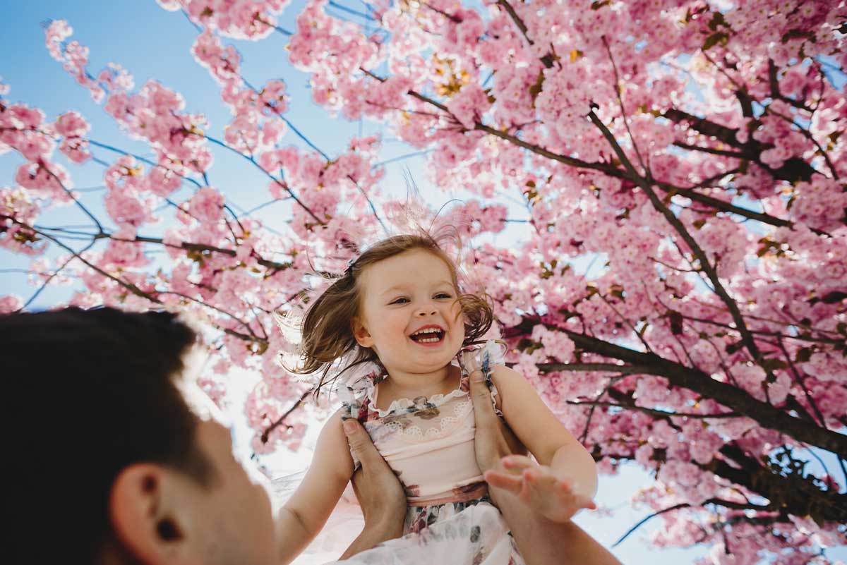 Dad holding his smiling baby girl up in the air outside with a cherry blossom tree behind.