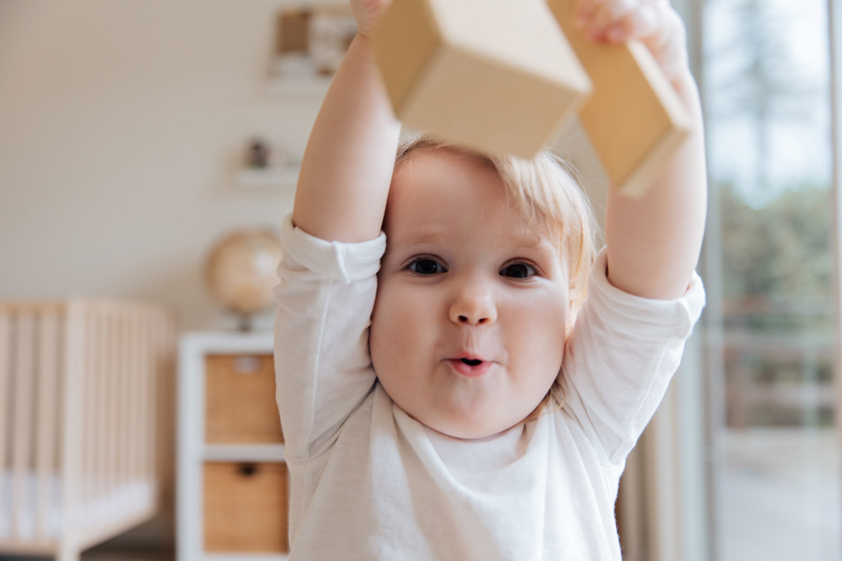A baby pulls a funny face at the camera as they lift both arms above their head.