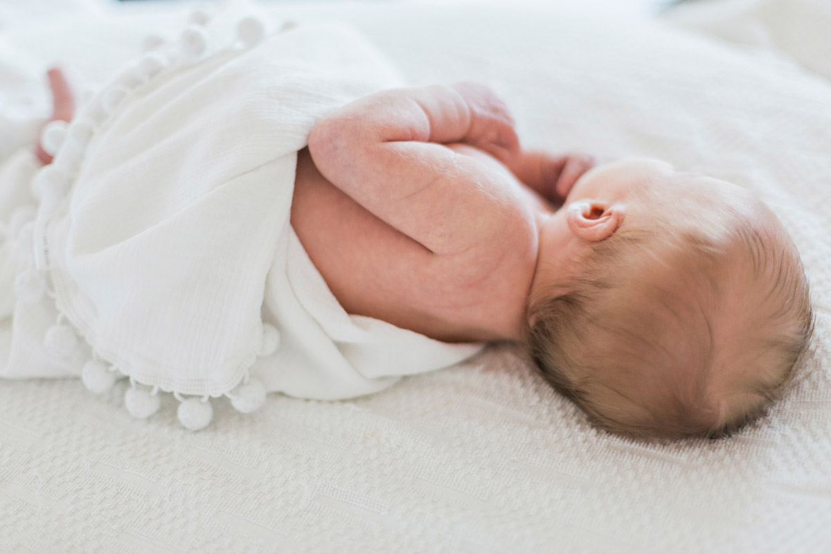 A newborn baby with blonde hair lies on a bed whilst wrapped in a white blanket.