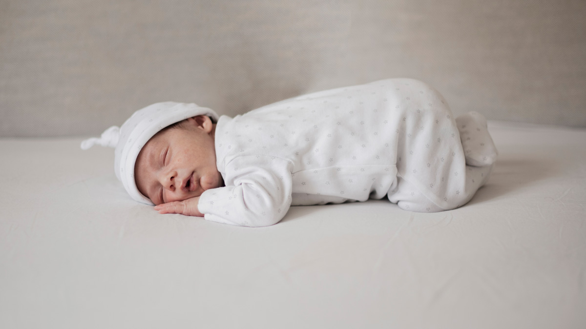 A newborn baby sleeping on a white mattress with a grey wall behind.