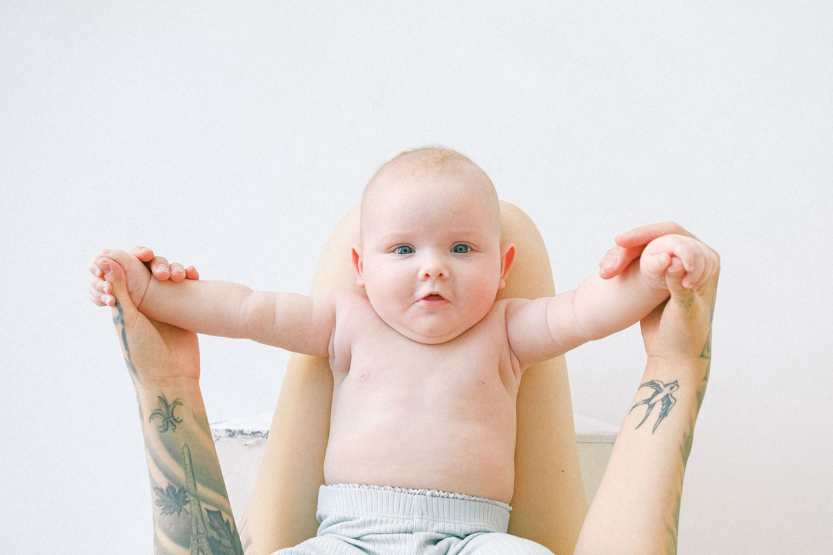A little baby lies on his back on his parent's lap holding their hands, he is looking up at the camera.