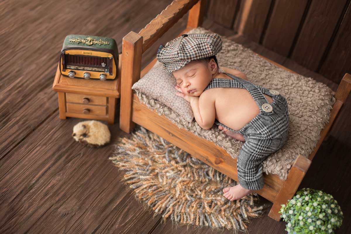 A baby boy is lying on his front, asleep on a miniature bed and wearing a checked hat.