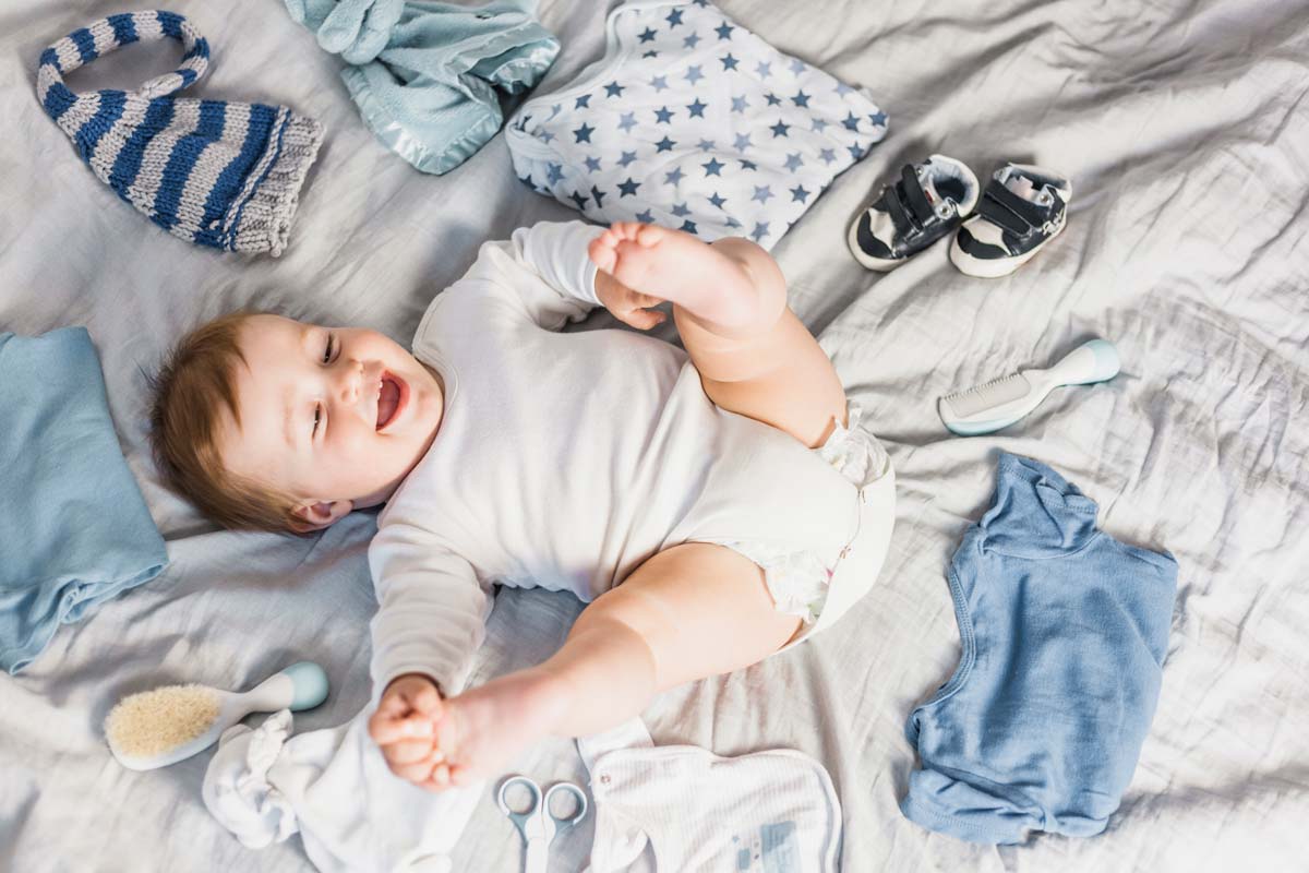 A baby boy is lying on a bed laughing, he is surrounded by lots of blue clothes.