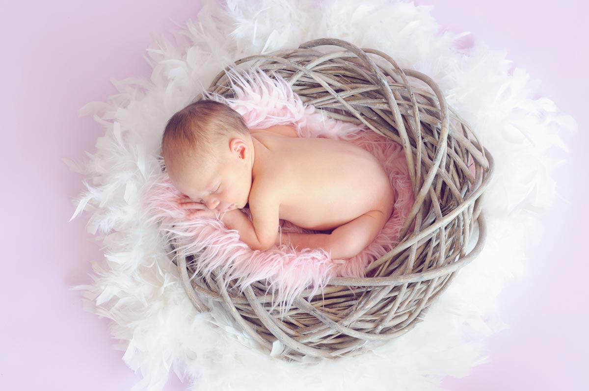 A baby girl is curled up asleep in a basket with a fluffy pink blanket.