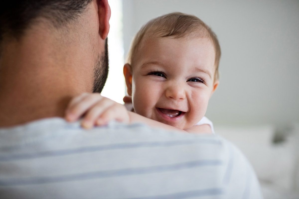 A baby girl is being held by her father, she is looking over his shoulder and laughing at the camera.