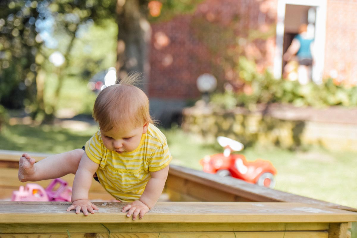 A brave baby girl climbs out of a large sandpit in the garden..