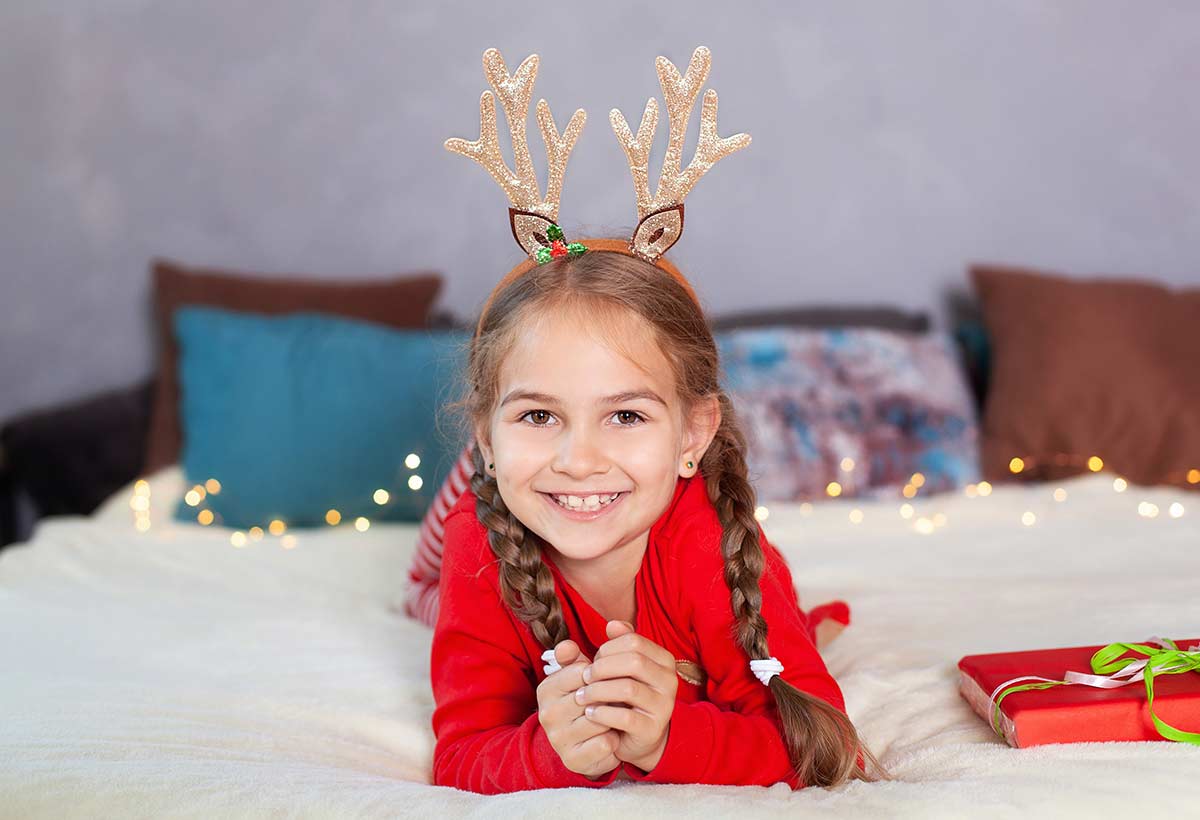 Little girl wearing gold deer antlers on her head, lying on the bed smiling.